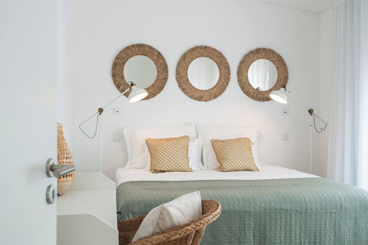 Une chambre à coucher avec un lit, trois miroirs fixés au mur, deux lampes de chevet, une chaise en osier et une porte blanche.