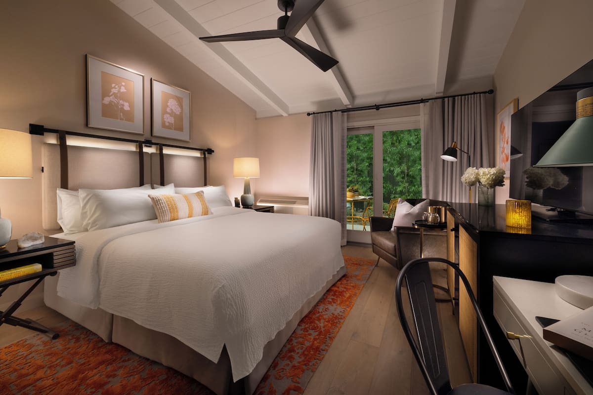 A hotel room with a large bed, bedside tables, a desk, and a chair. There are two framed pictures on the wall, a ceiling fan, and a window with curtains.