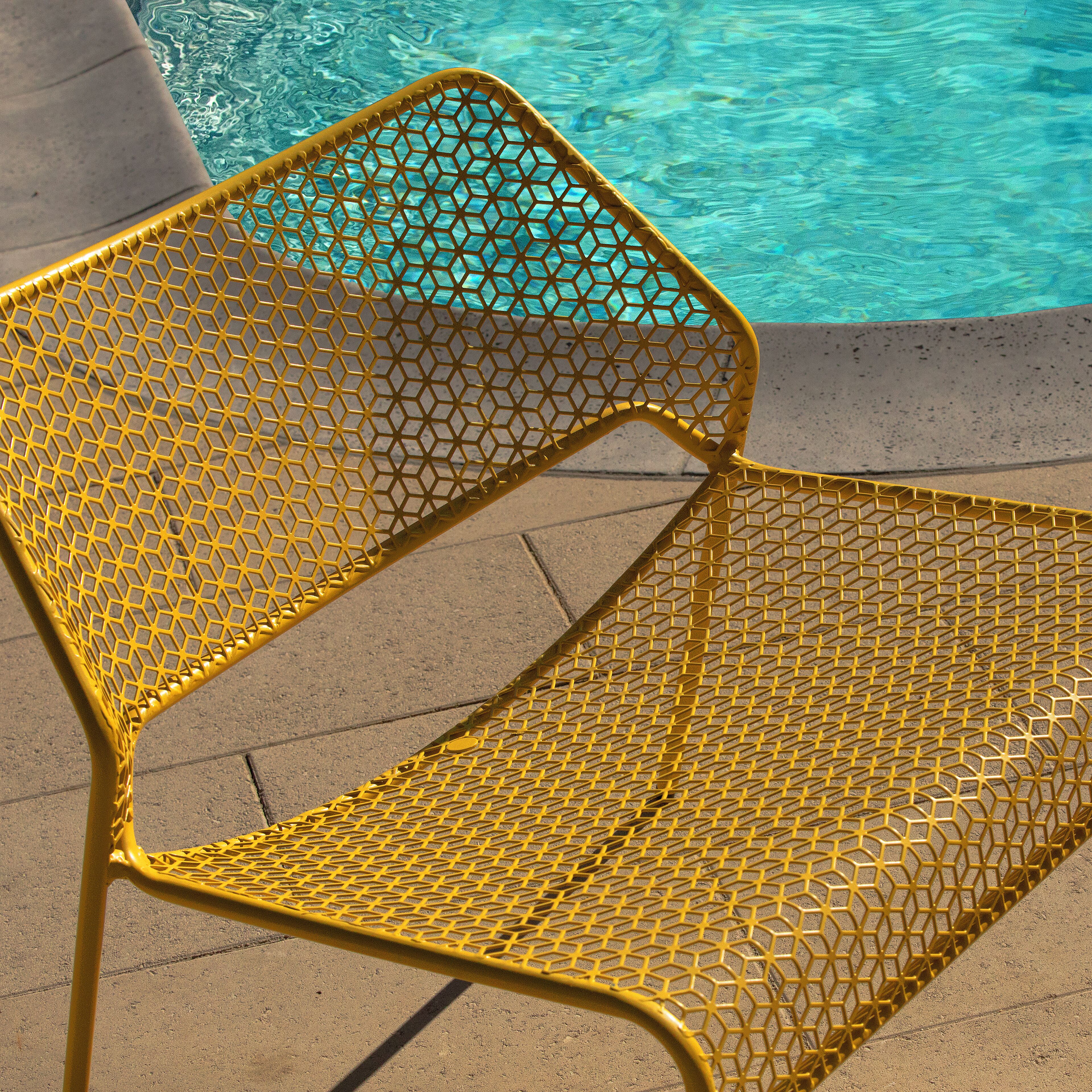 A yellow metal chair with a geometric pattern, positioned on a concrete surface near a swimming pool.
