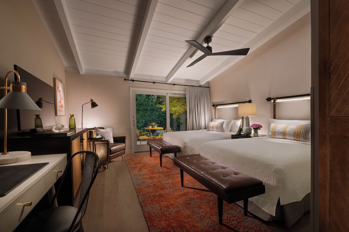 A hotel room with a large bed, a desk with a lamp, a chair, a bench, a ceiling fan, and a view of the outdoors.