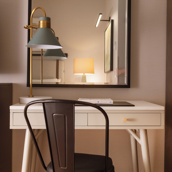 Quarto de hotel com uma escrivaninha branca, uma cadeira, uma luminária de mesa, uma luminária de cabeceira e um espelho.