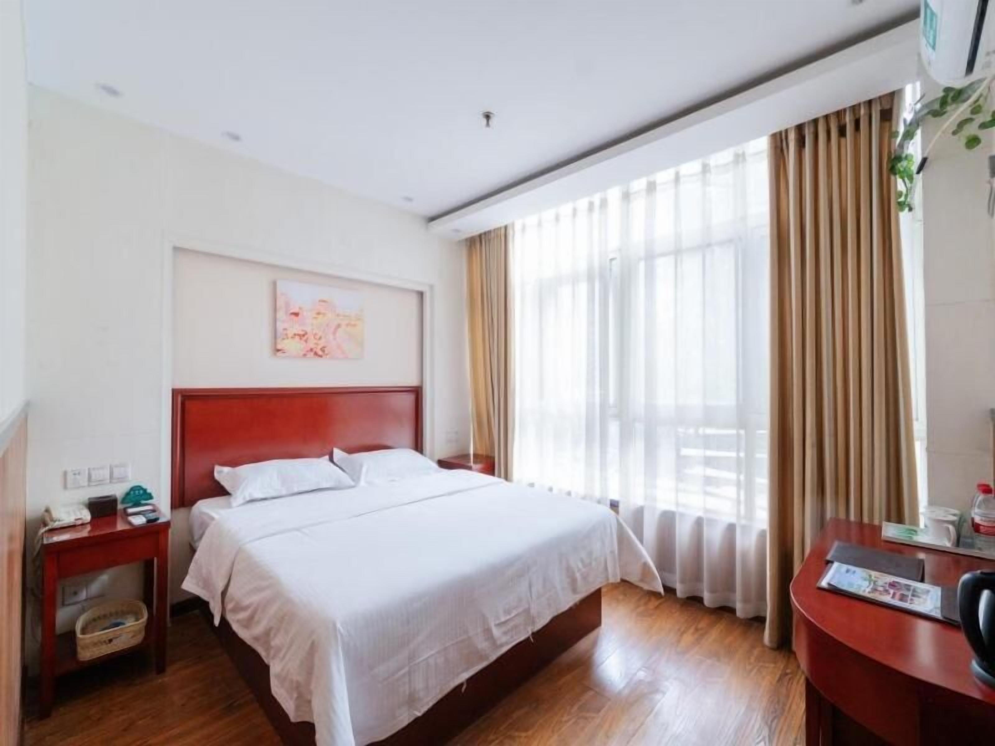 Ein Hotelzimmer mit einem großen Bett, einem Holzkopfende, einem Nachttisch mit Lampe, einem Schreibtisch mit Stuhl und einem fenster mit Vorhängen.