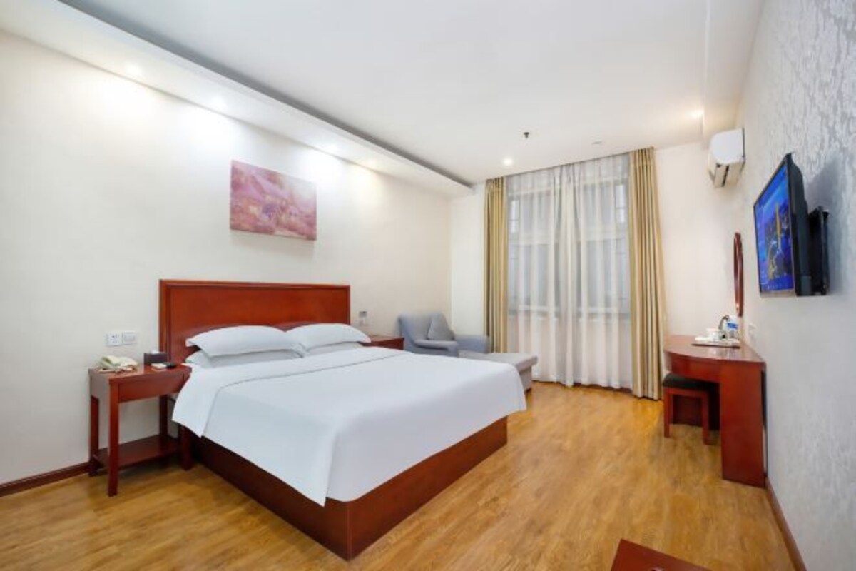 Une chambre d’hôtel avec un lit en bois, un bureau, une chaise, une télévision et un tableau au mur.