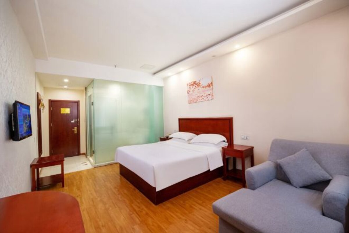 Une chambre d’hôtel avec un lit, un canapé gris, un banc en bois, une télévision et une porte donnant sur une autre pièce.
