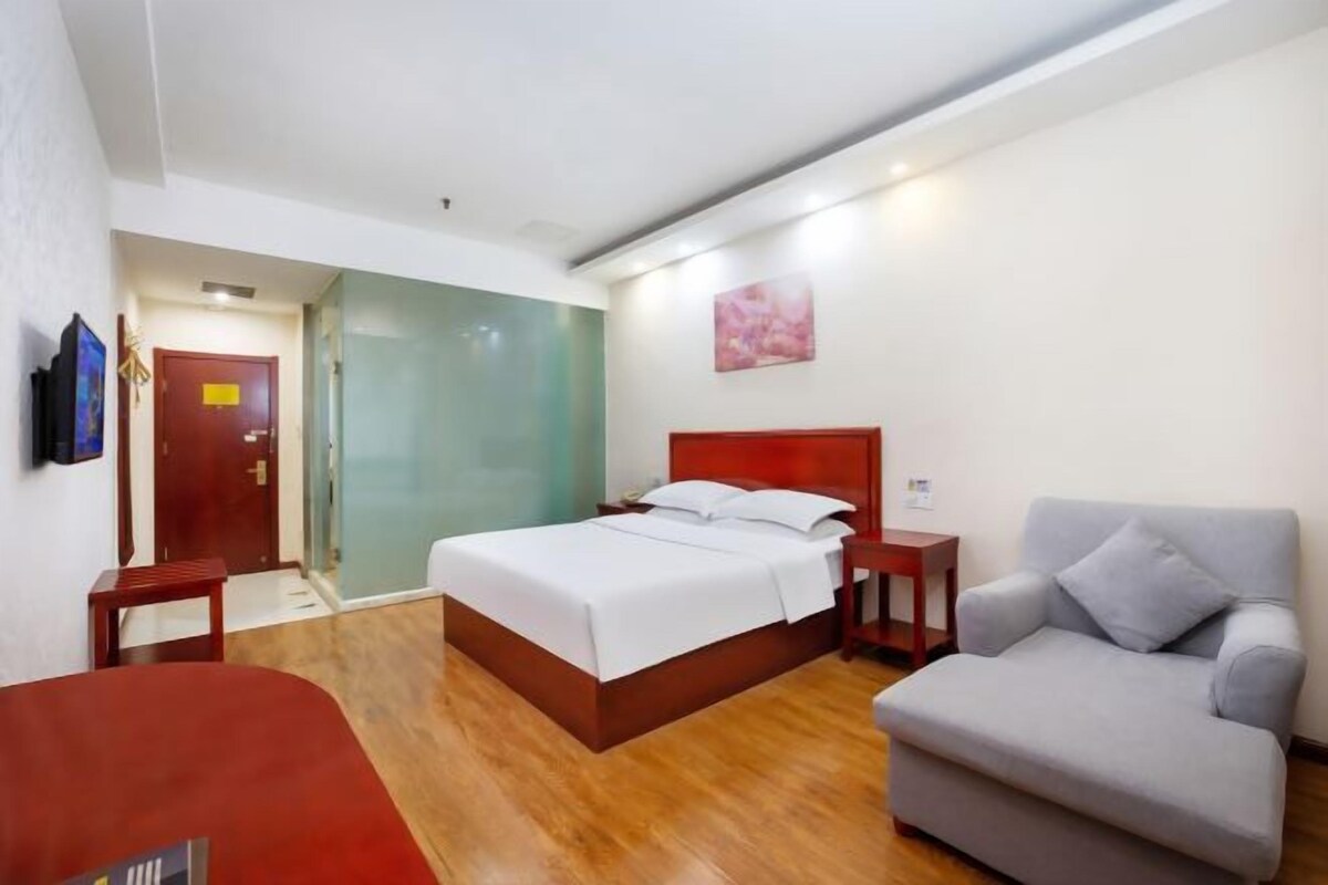 A hotel room with a bed, a grey armchair, a wooden floor, a red door, and a glass partition.
