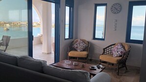 A room with a view of the sea, a sofa, two chairs, a table, and a clock on the wall.