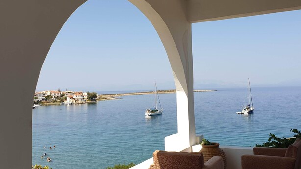 A seaside view from a covered terrace with two sailboats on the water, a coastal village in the background, and a seating area with cushions and a potted plant.