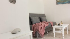 Un salon minimaliste avec un canapé gris, une table basse blanche et une console blanche. Il y a deux coussins sur le canapé et un plaid rose.