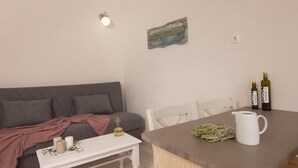 Un salon chaleureux avec un canapé gris, une petite table blanche ornée de bougies et d’une bouteille, un tableau accroché au mur, et un coin repas avec une chaise blanche et deux bouteilles d’huile d’olive.