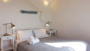 A neatly made bed with white linens and a rolled towel, two bedside lamps, a wall-mounted light fixture, and a hanging light fixture above the bed.