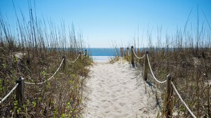 Un sentier de sable qui longe la plage et mène à l’océan, bordé de hautes herbes et de poteaux en bois.