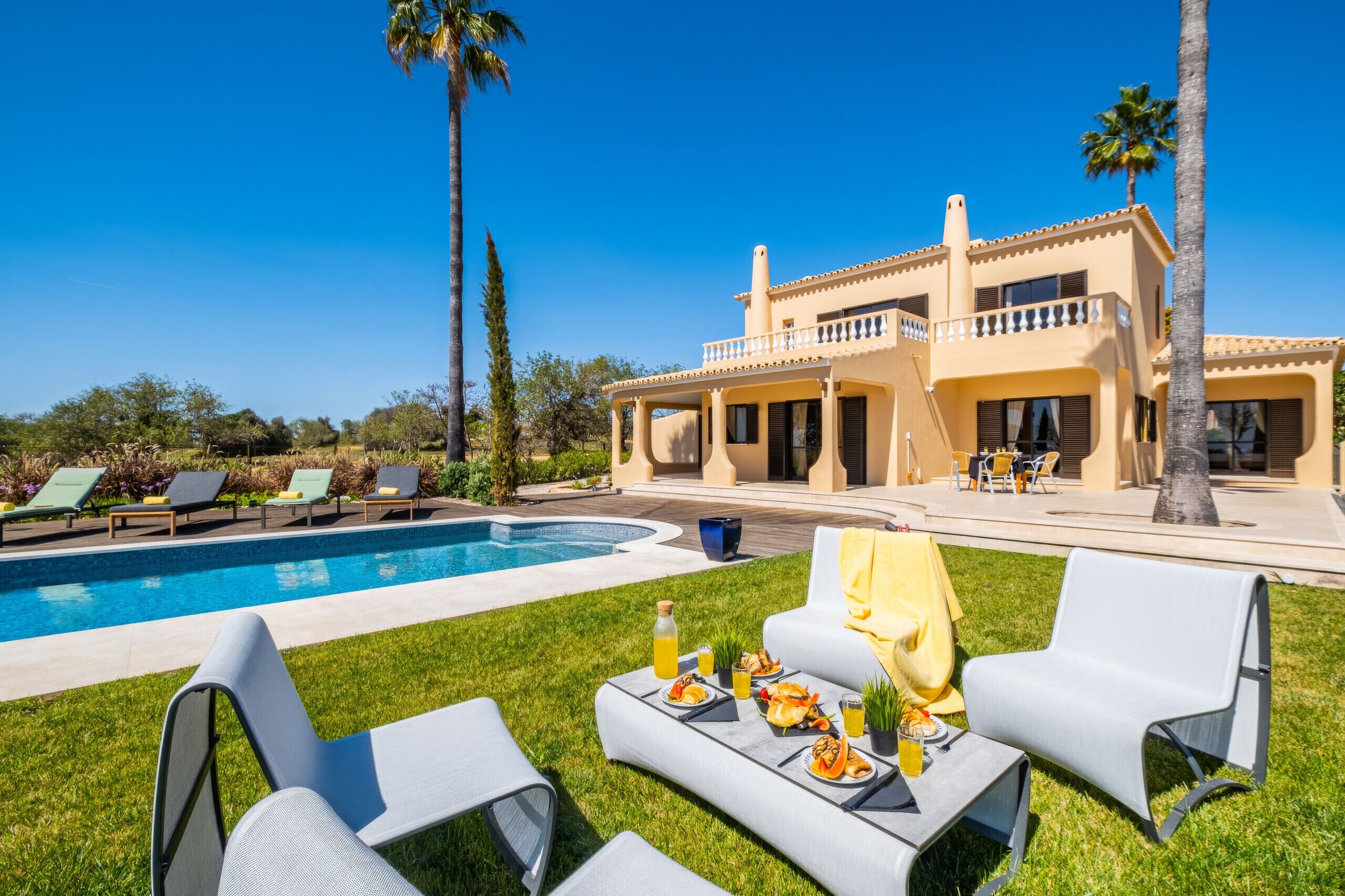 Un espace aménagé au bord de la piscine avec des chaises longues, une table à manger avec de la nourriture et des boissons, et une grande maison dotée d’un balcon.