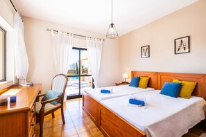 Habitación de hotel con dos camas, un escritorio, una silla, una ventana con cortinas, una lámpara colgante y un cuadro en la pared.