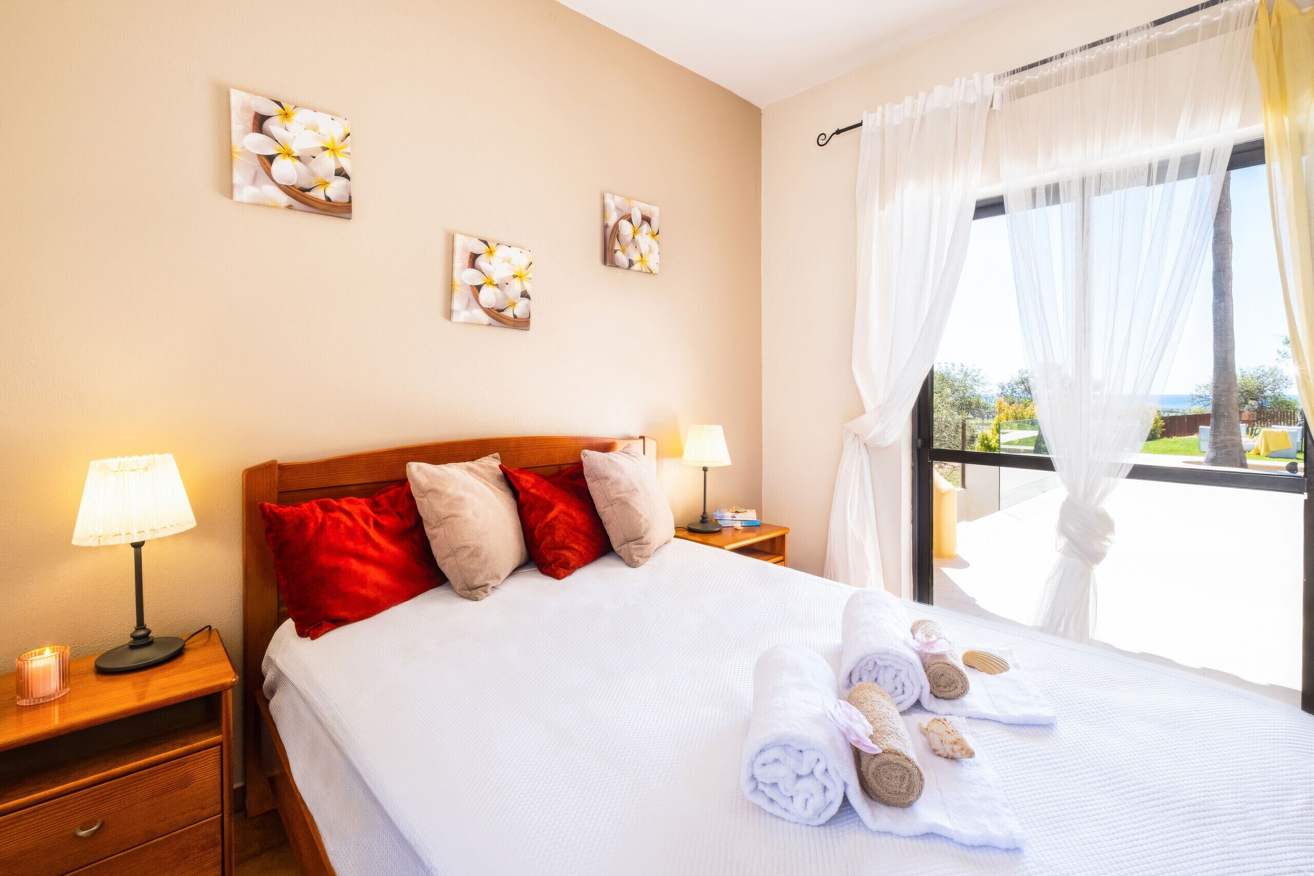 A hotel room with a bed, pillows, a nightstand, a lamp, and a window with sheer curtains.