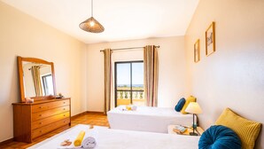 Une chambre d’hôtel avec deux lits, une commode en bois, un miroir, une fenêtre avec des rideaux, un balcon donnant sur une vue agréable et une table de chevet avec une lampe.