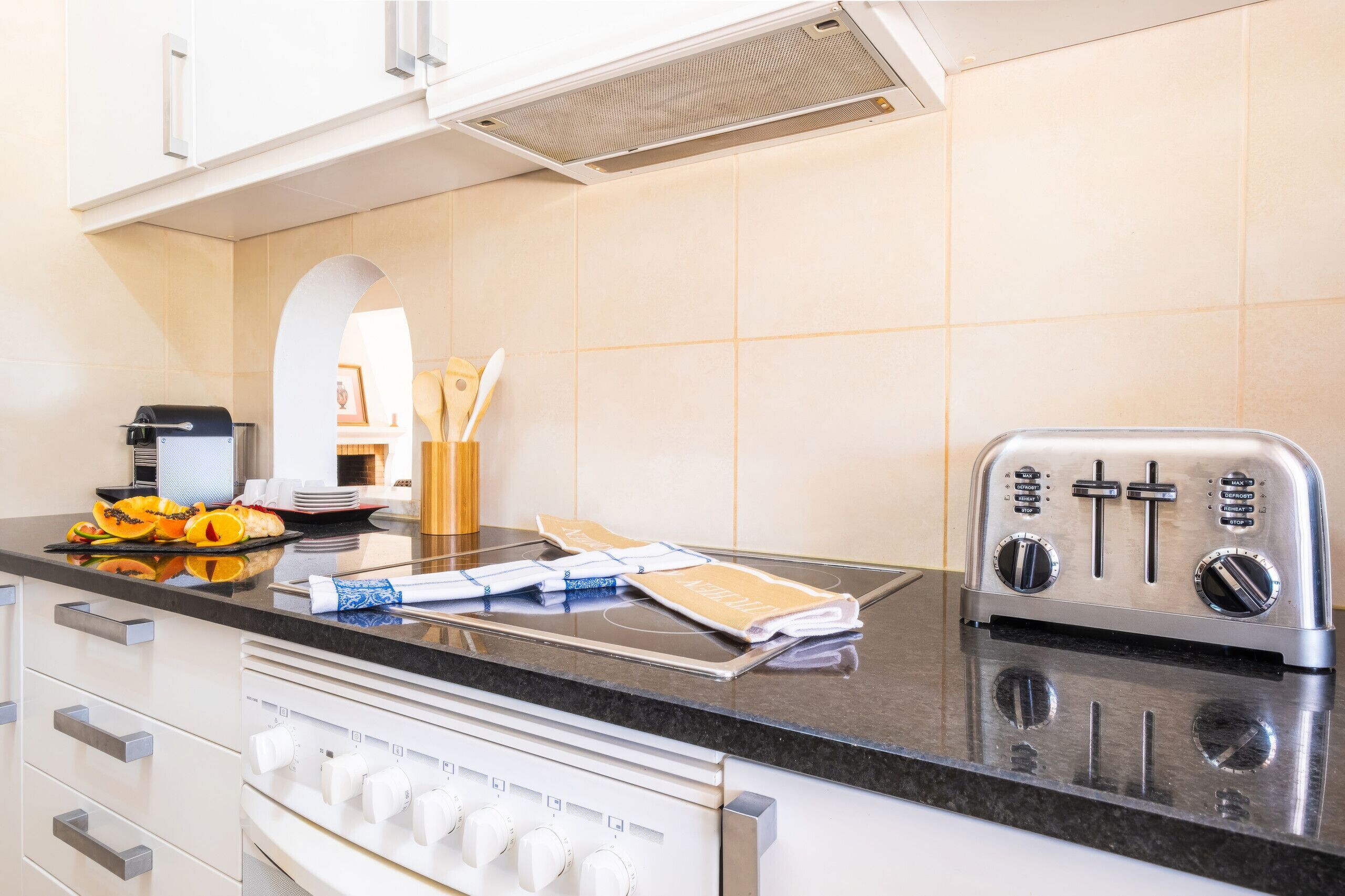 Une cuisine moderne équipée d’un grille-pain encastré, d’une cuisinière dotée de plusieurs commandes et d’un plan de travail agrémenté d’une planche à découper, d’ustensiles de cuisine et d’une assiette de fruits.