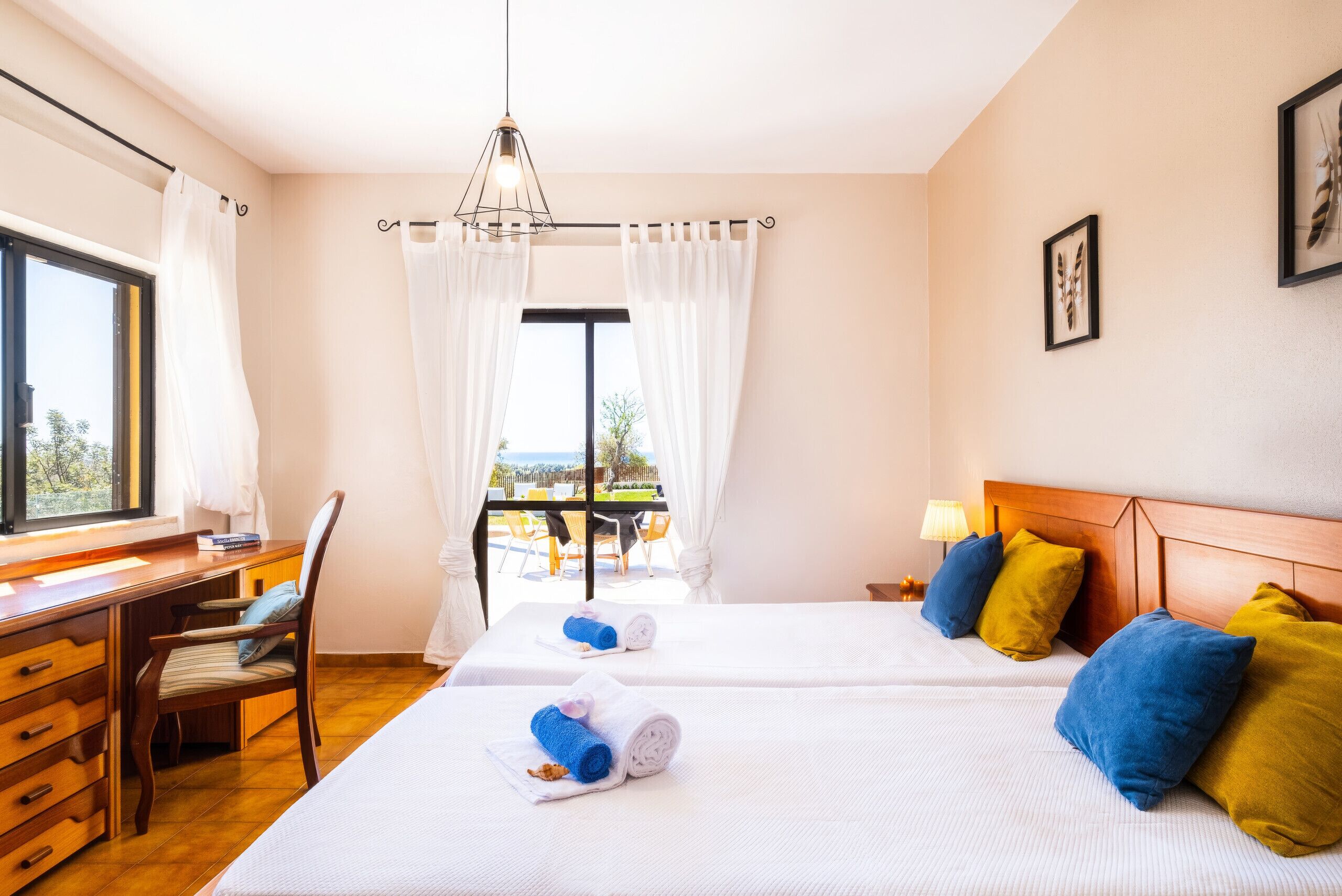 A hotel room with a desk, two beds, a window with curtains, a view of the outdoors, and a chandelier.