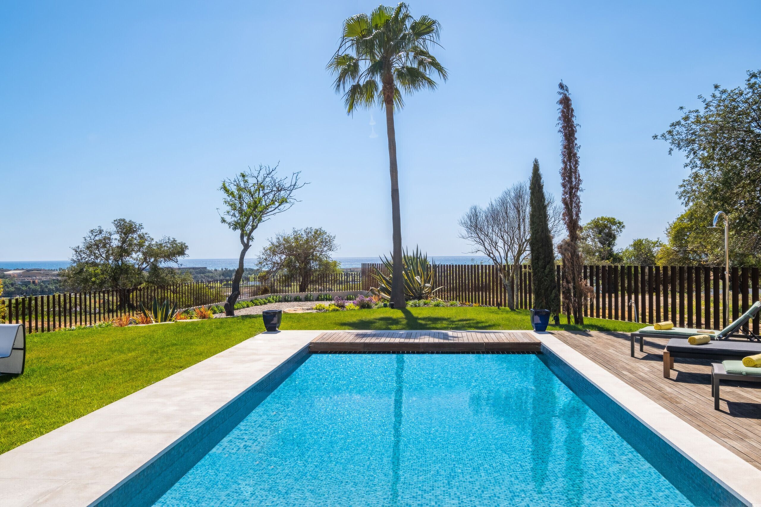 Une piscine rectangulaire dotée d’une terrasse en bois et de chaises longues, entourée d’une clôture en bois et de diverses plantes.