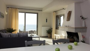 Un salón espacioso con sofá, mesa de comedor y sillas. Se ve el mar a través de amplias ventanas. En el fondo, se distingue una chimenea.