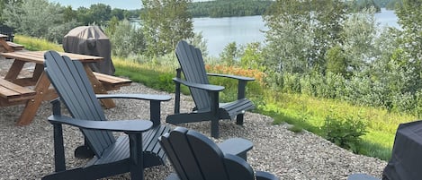 Espace extérieur aménagé avec des chaises Adirondack et des tables de pique-nique, offrant une vue sur un lac bordé d’arbres et entouré de collines.