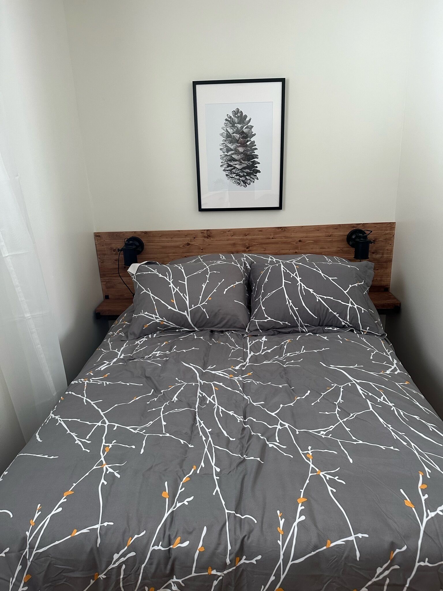 Un lit avec un motif de branches grises et blanches, une œuvre d’art représentant une pomme de pin encadrée au-dessus, et une tête de lit en bois.
