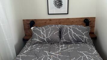 Un lit avec un motif de branches grises et blanches, une œuvre d’art représentant une pomme de pin encadrée au-dessus, et une tête de lit en bois.