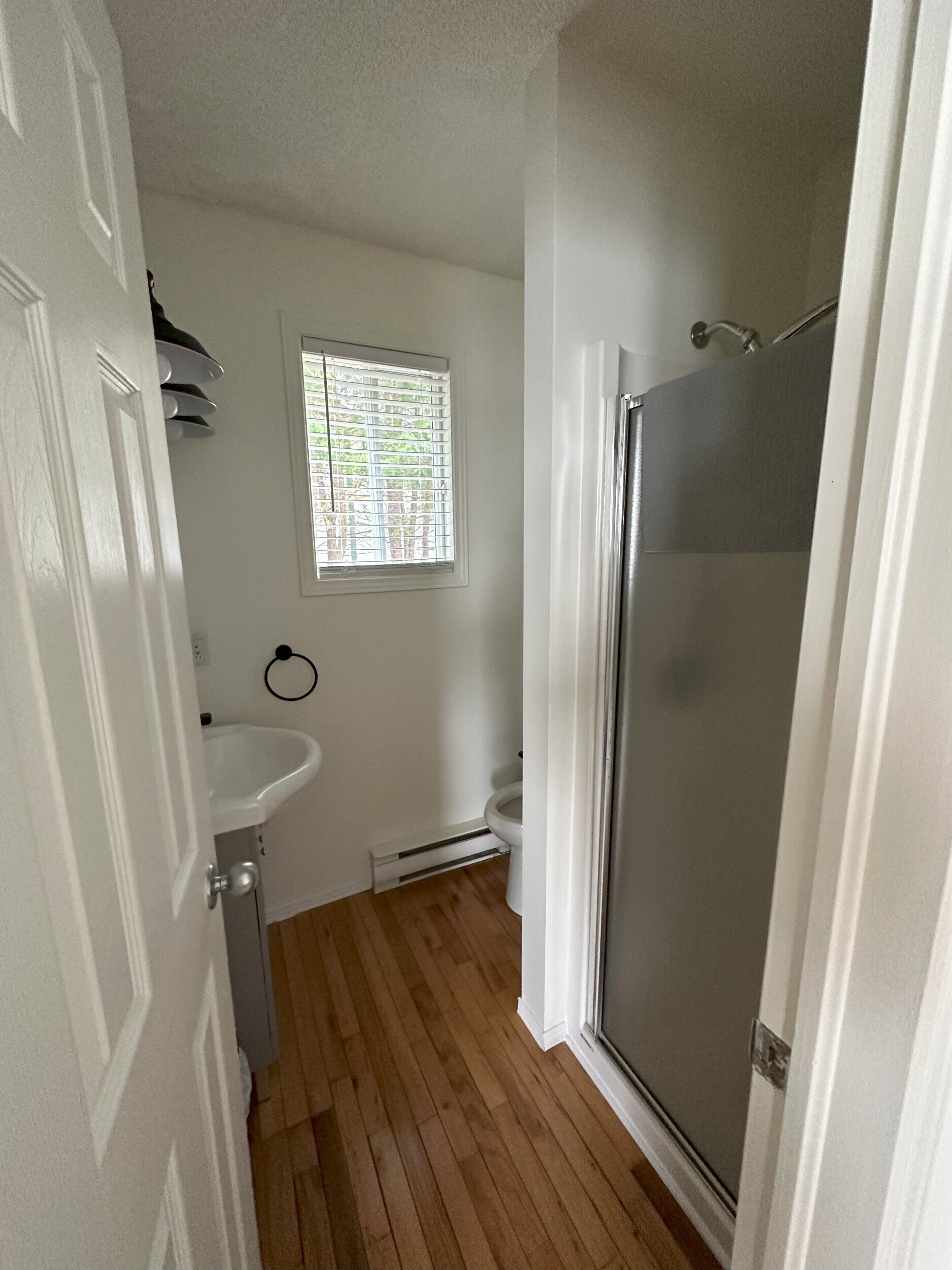 A bathroom with a shower, toilet, sink, and a window with blinds.