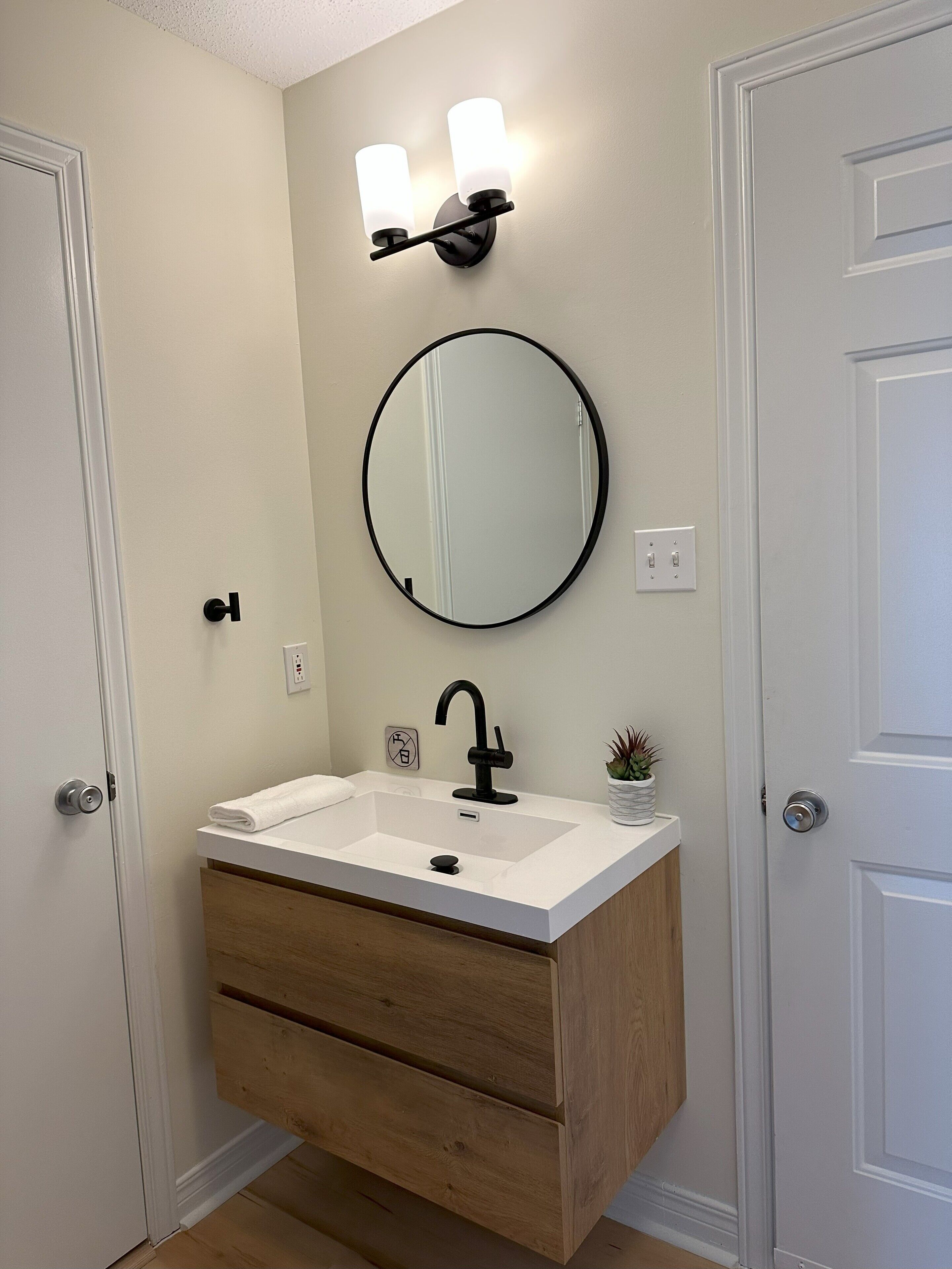 A bathroom with a white sink, black faucet, and a round mirror. There are two wall-mounted lights above the mirror and a small potted plant on the sink's edge.