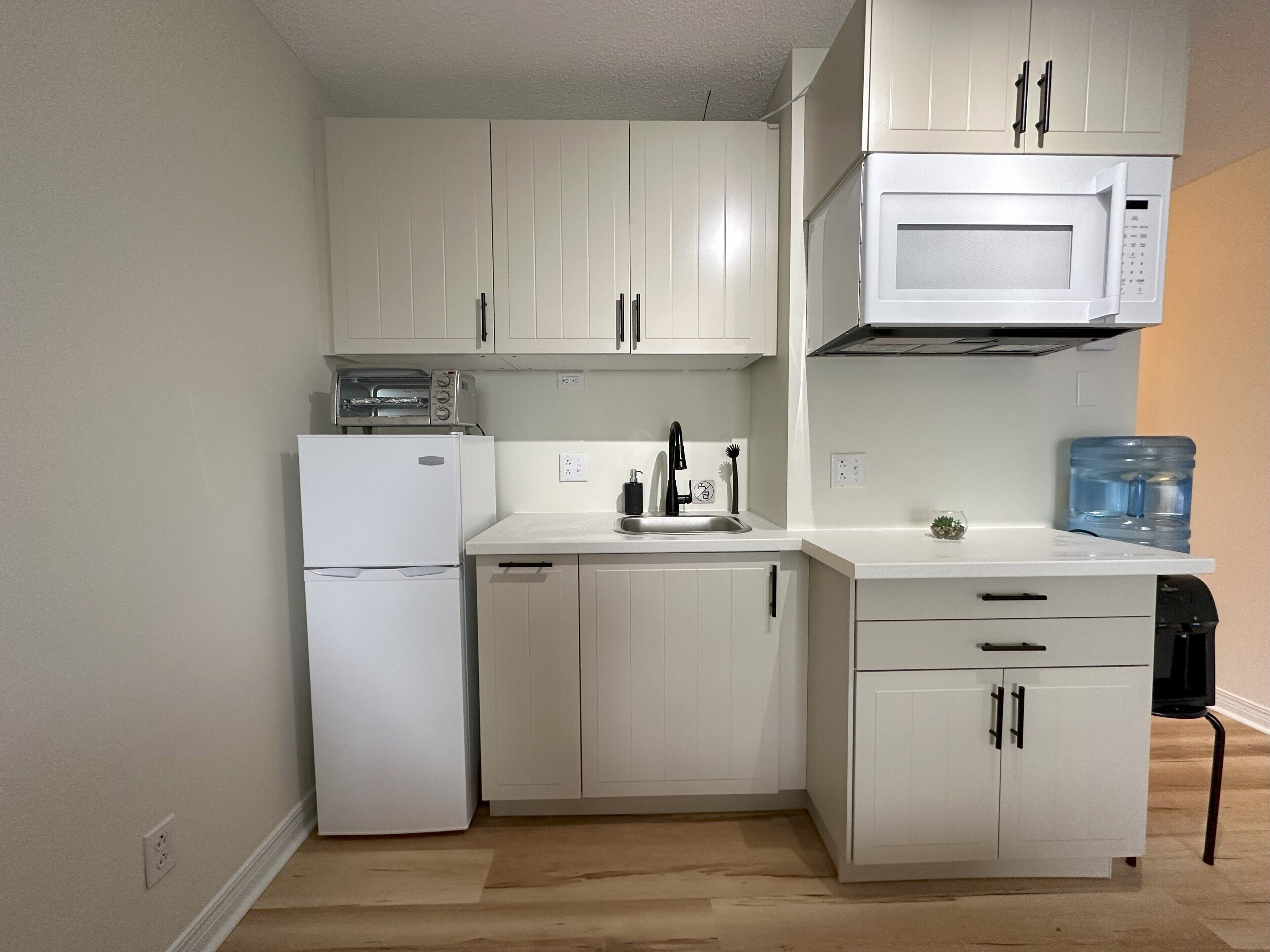 Cocina compacta con gabinetes blancos, microondas, tostadora, lavabo y dispensador de agua.