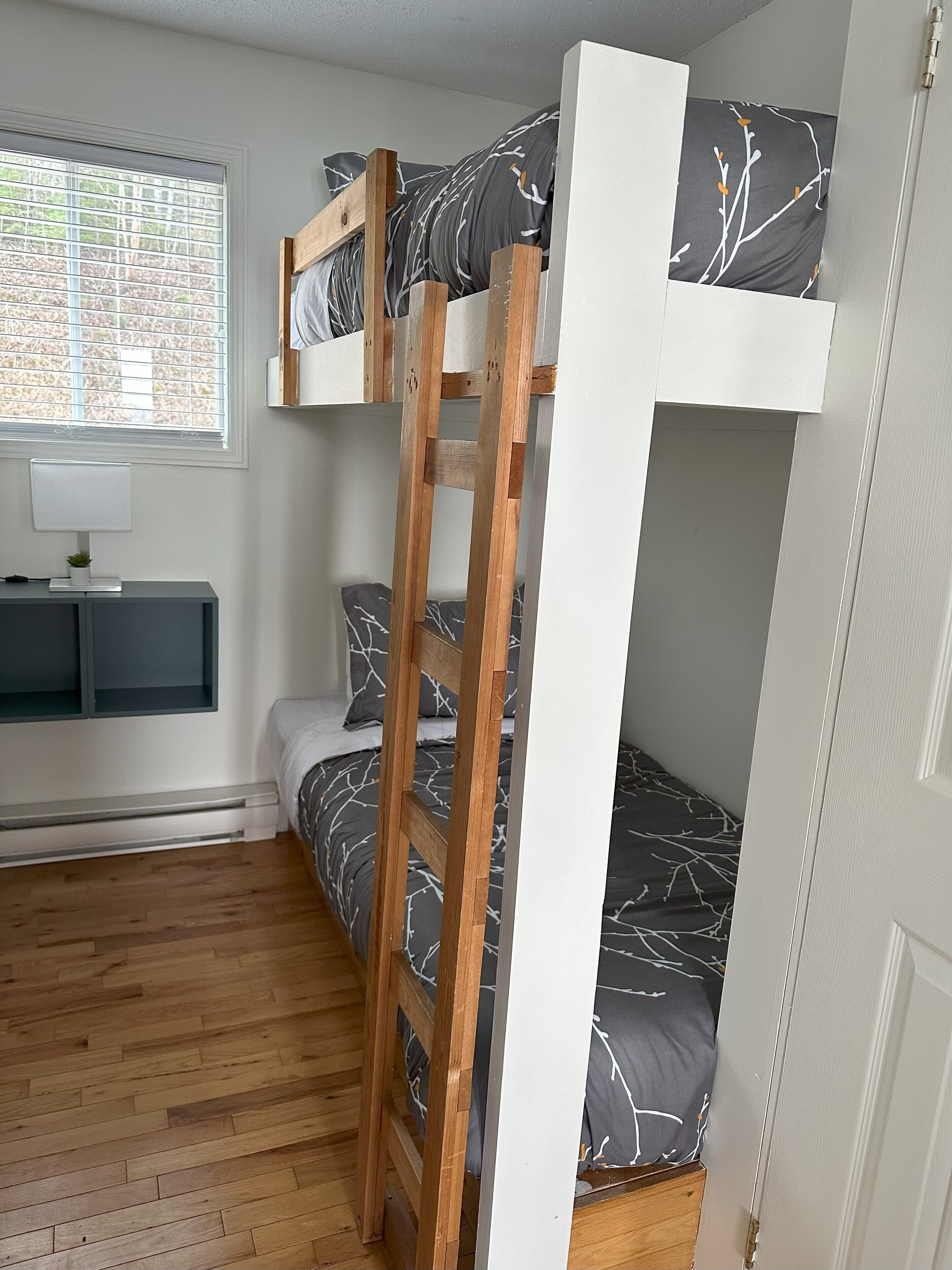A bunk bed with a ladder, a bedside table with a lamp, and a window with blinds.