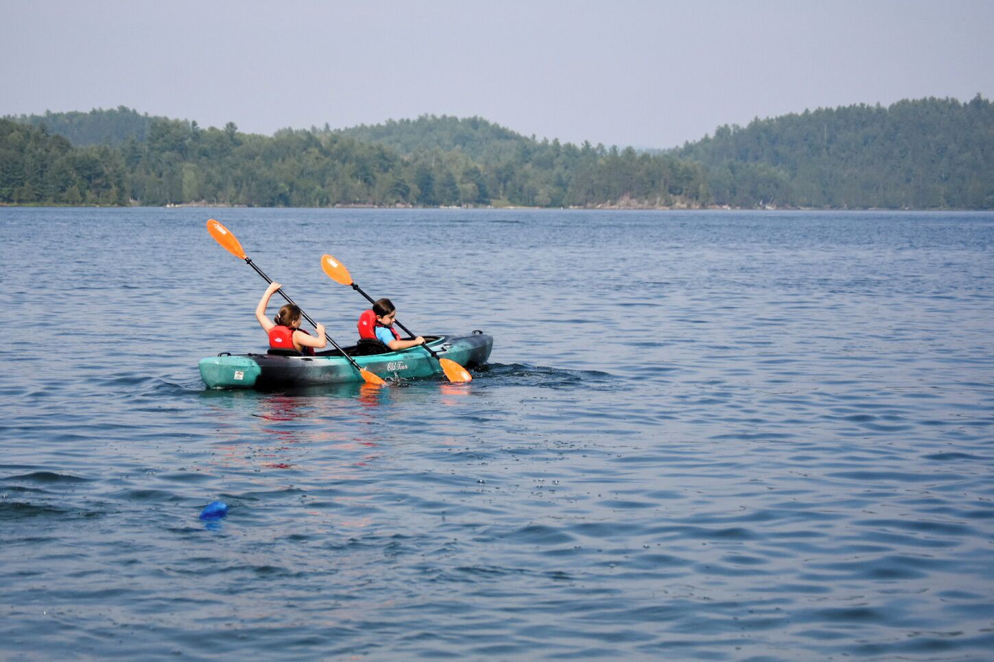 Two people kayaking on a calm lake with a forested shoreline in the background.