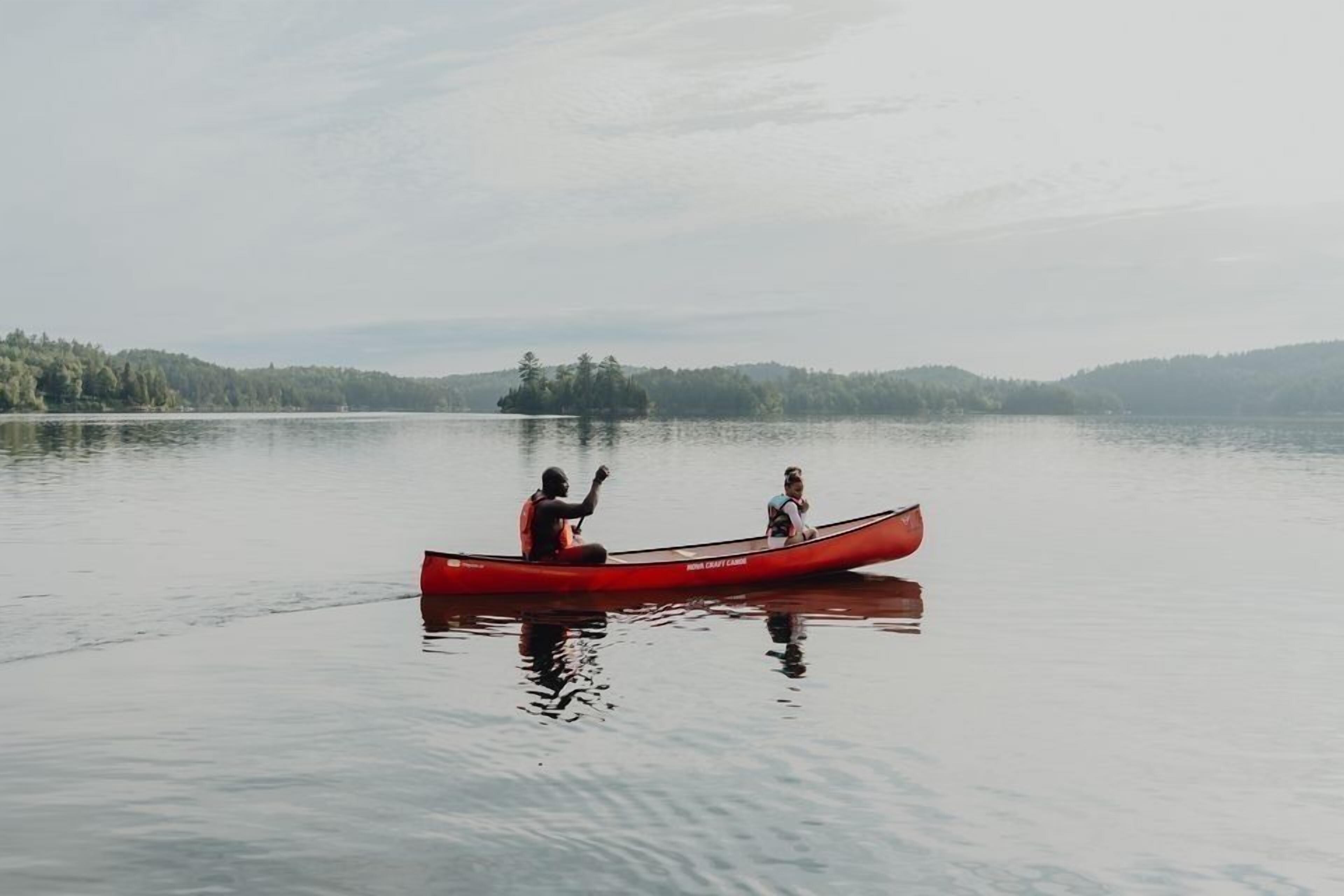 A red canoe on calm water with two people, surrounded by a forested landscape.