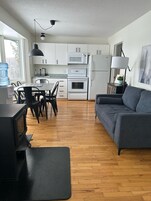 Una cocina moderna con armarios blancos, un microondas, horno y refrigerador. Hay una mesa de comedor con sillas, un sofá gris y un mueble negro para televisión.