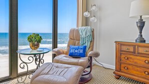 A room with a view of the ocean, a leather chair with a beach-themed pillow, a small table with a plant, and a wooden dresser with a lamp.