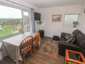 A living room with a dining table, chairs, a sofa, a TV, and a window with a view of a green landscape.