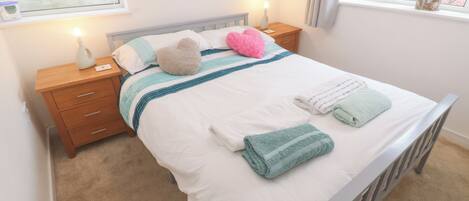 Una cama bien hecha con edredón blanco, dos almohadas, una manta a rayas verde azulada y blanca, y dos toallas dobladas. Hay una mesita de noche de madera con una lámpara y un pequeño objeto decorativo junto a la cama.