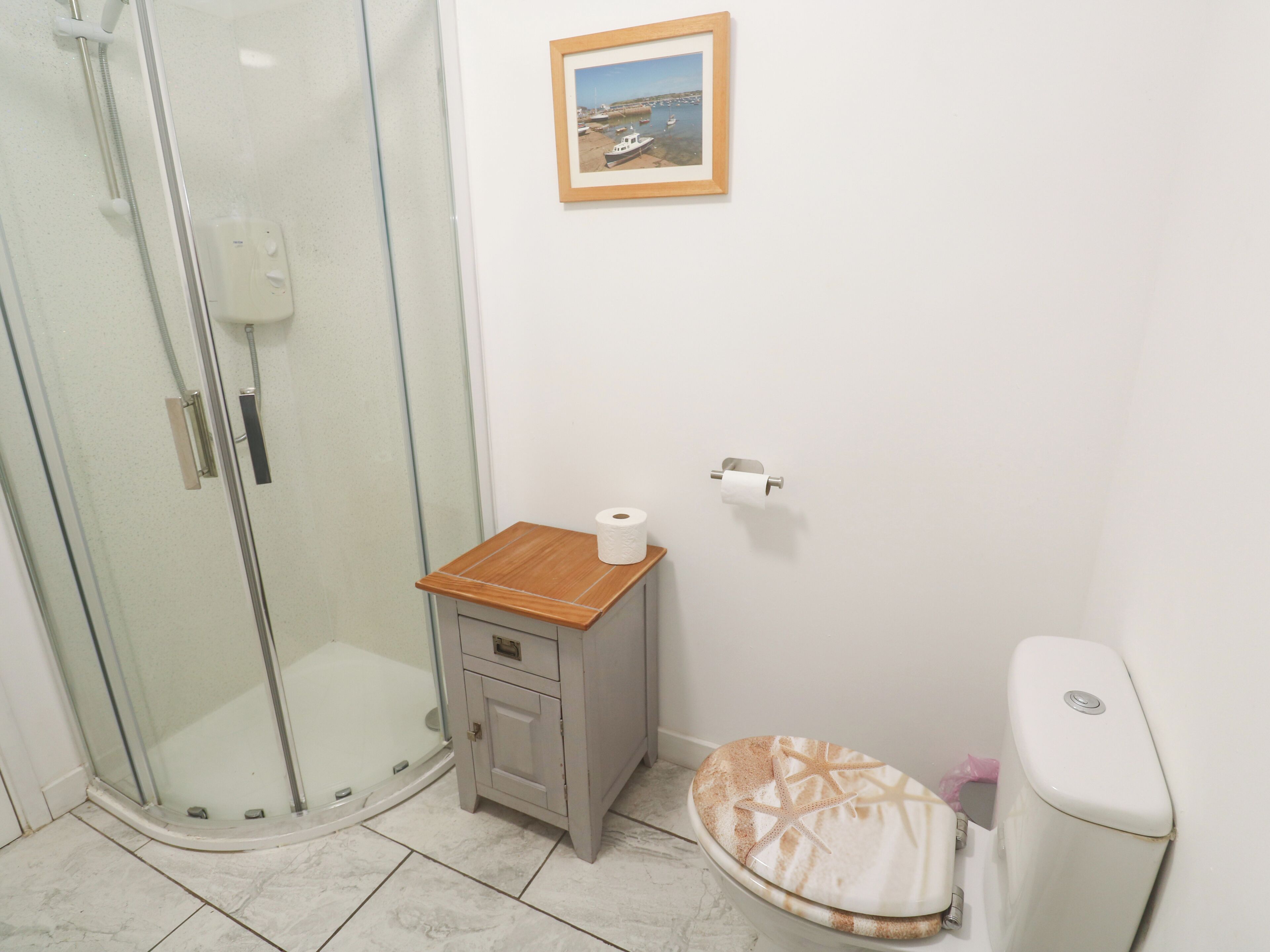 Une salle de bain avec une douche en verre, un petit meuble en bois, des toilettes décorées de motifs en forme d’étoile de mer et un tableau encadré au mur.
