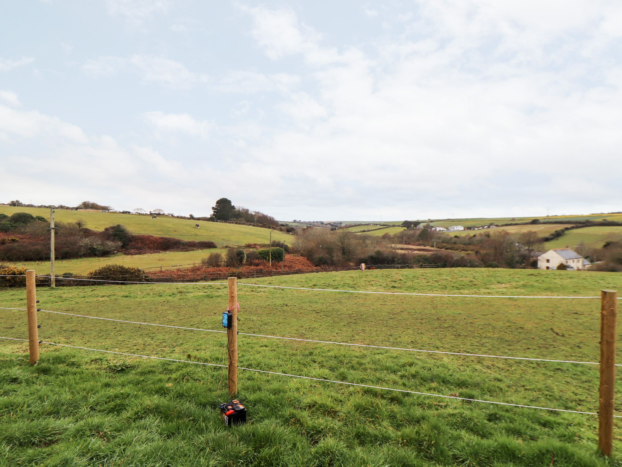 A rural landscape with a fenced field, a wooden post with a device, and a distant house.
