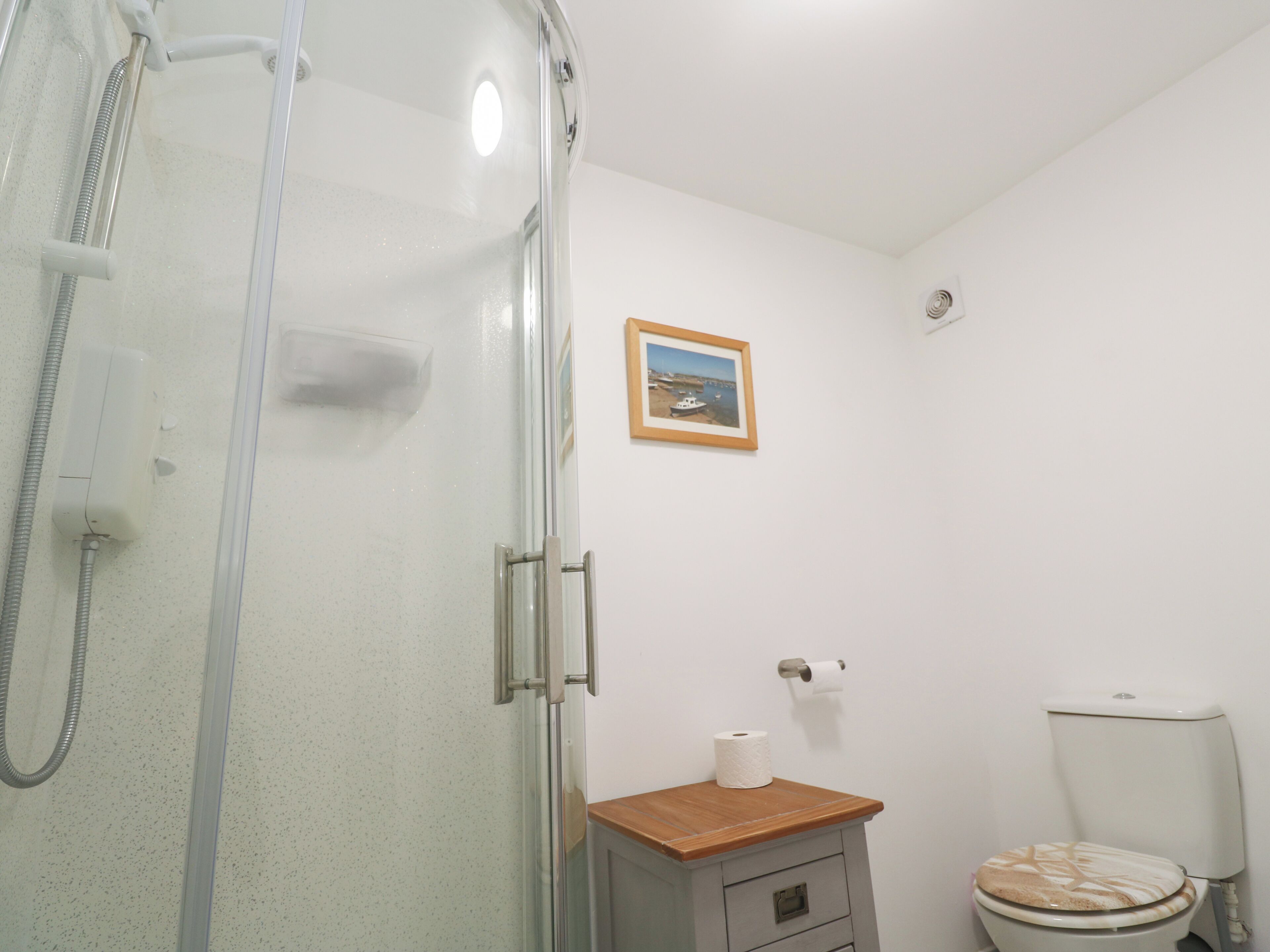 A bathroom with a glass shower enclosure, a toilet, a small wooden cabinet, and a framed picture on the wall.