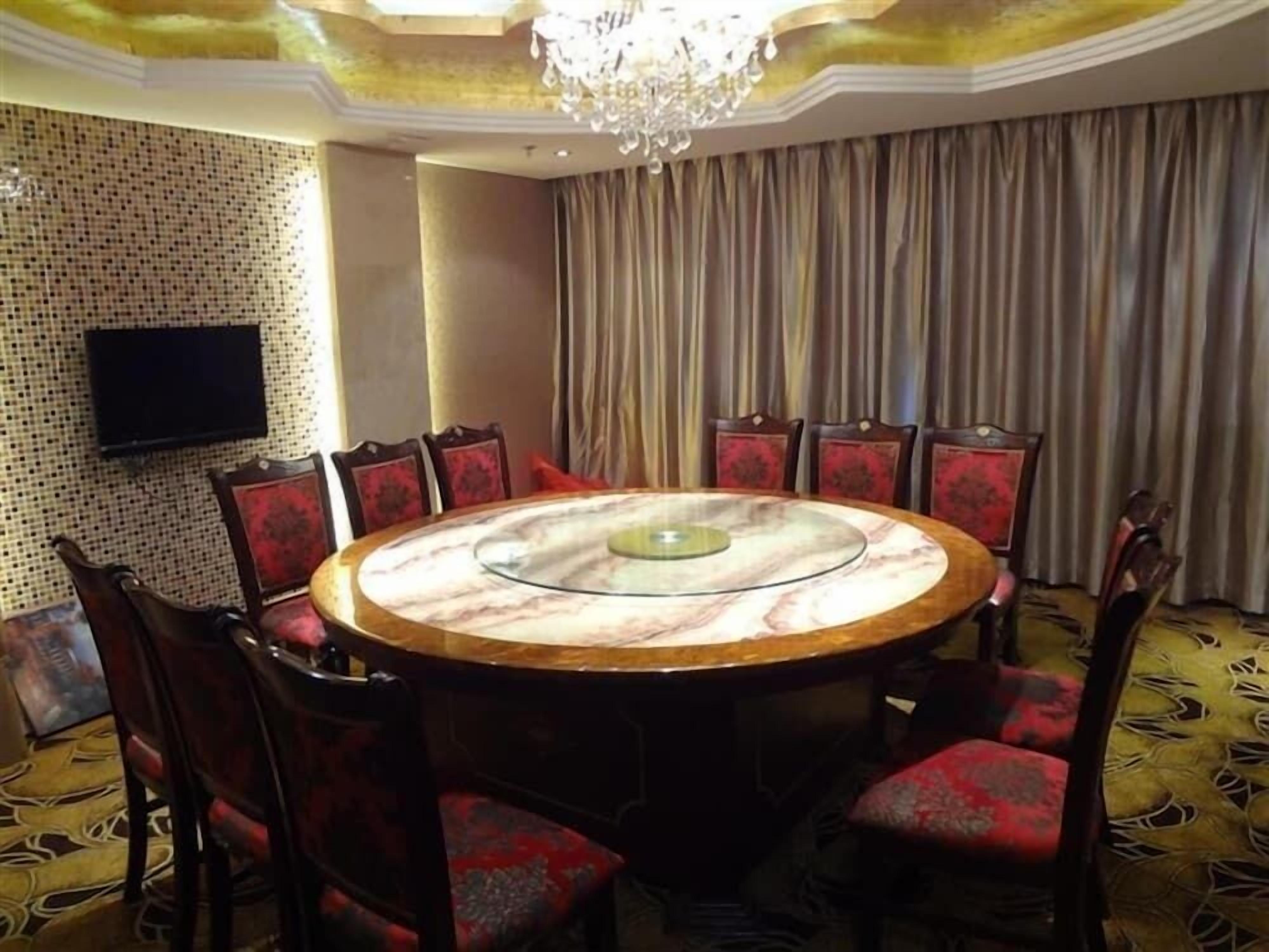 A round table with a glass top surrounded by chairs with red cushions, a chandelier, a television on the wall, and a patterned carpet.