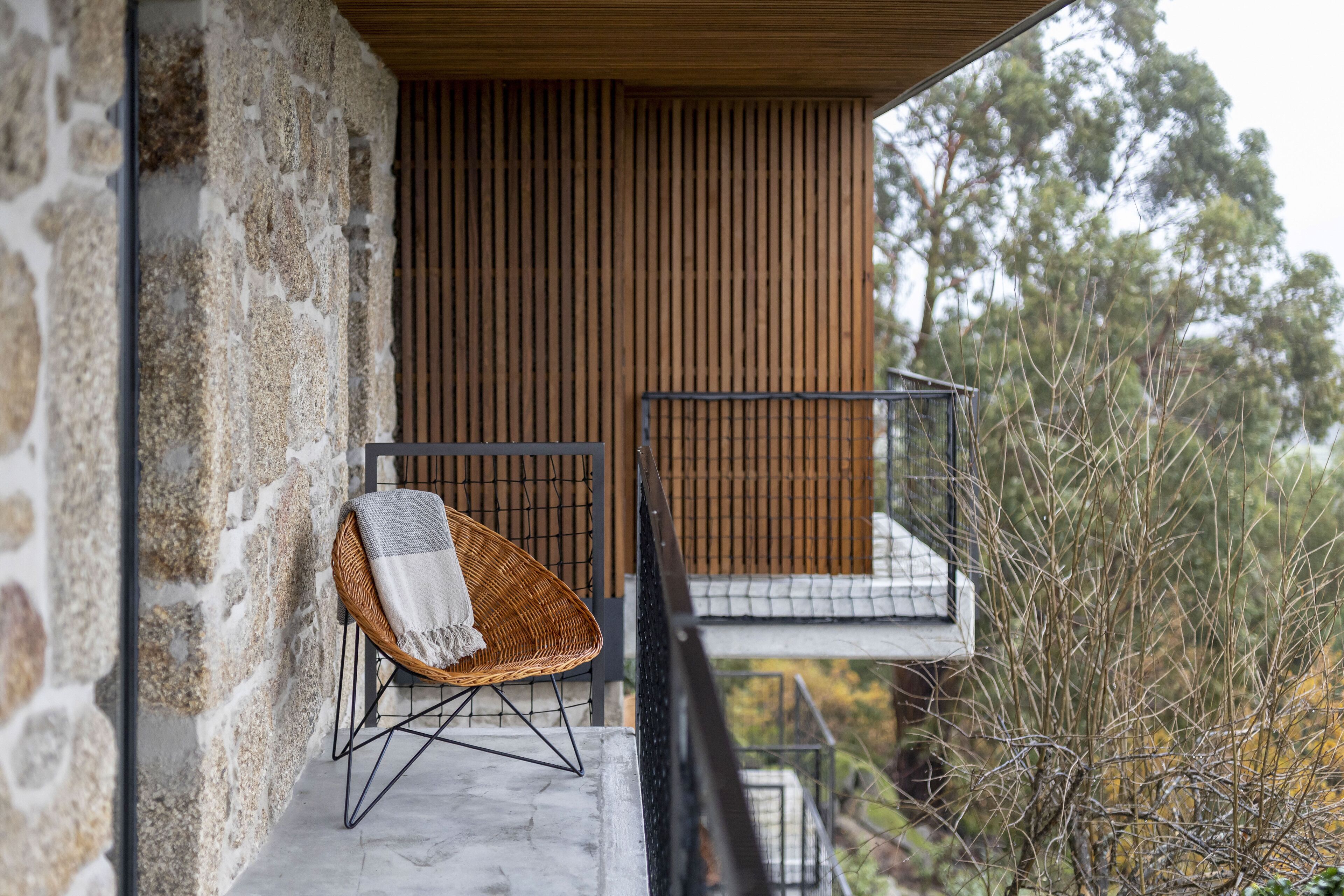Pared exterior de piedra, una silla de mimbre con una manta y barandilla de balcón de listones de madera.