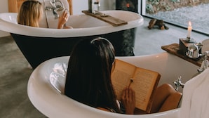 A modern bathroom with two freestanding tubs, a wooden shelf with toiletries, and a large window with a green ivy wall. - TheVagar  (Belmonte)