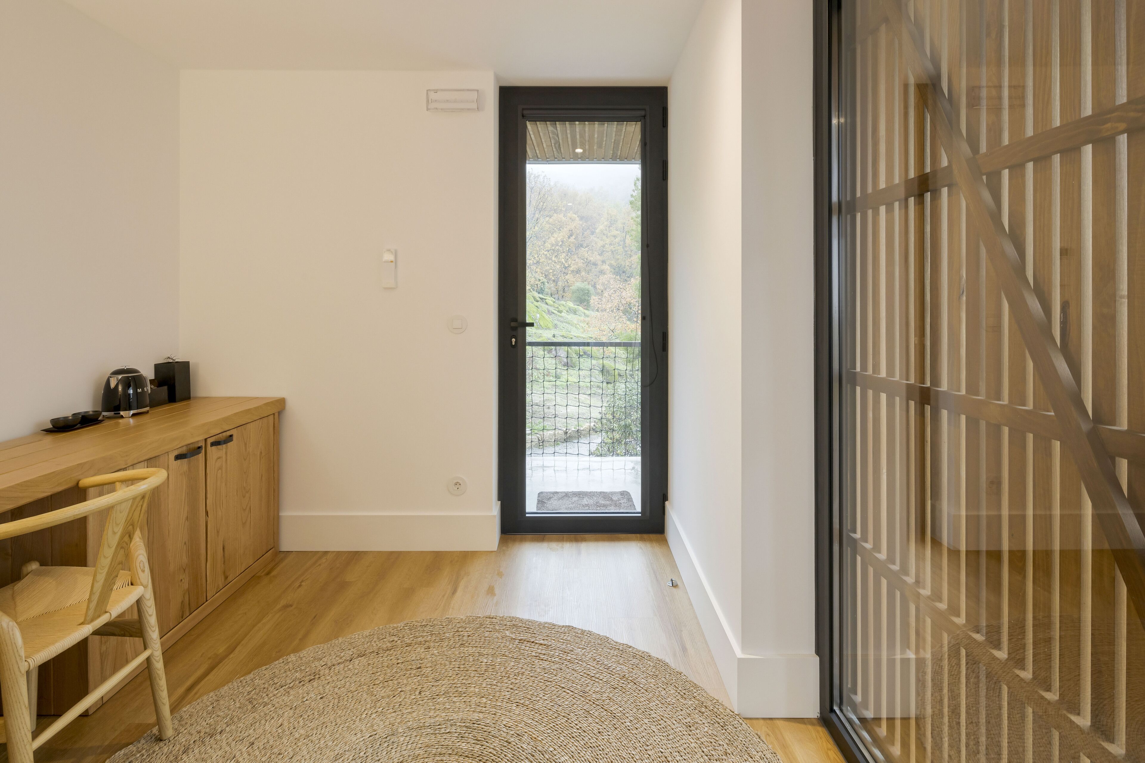 Une chambre moderne avec un bureau en bois, une chaise, un petit tapis et une vue sur un balcon extérieur, accessible par une porte.