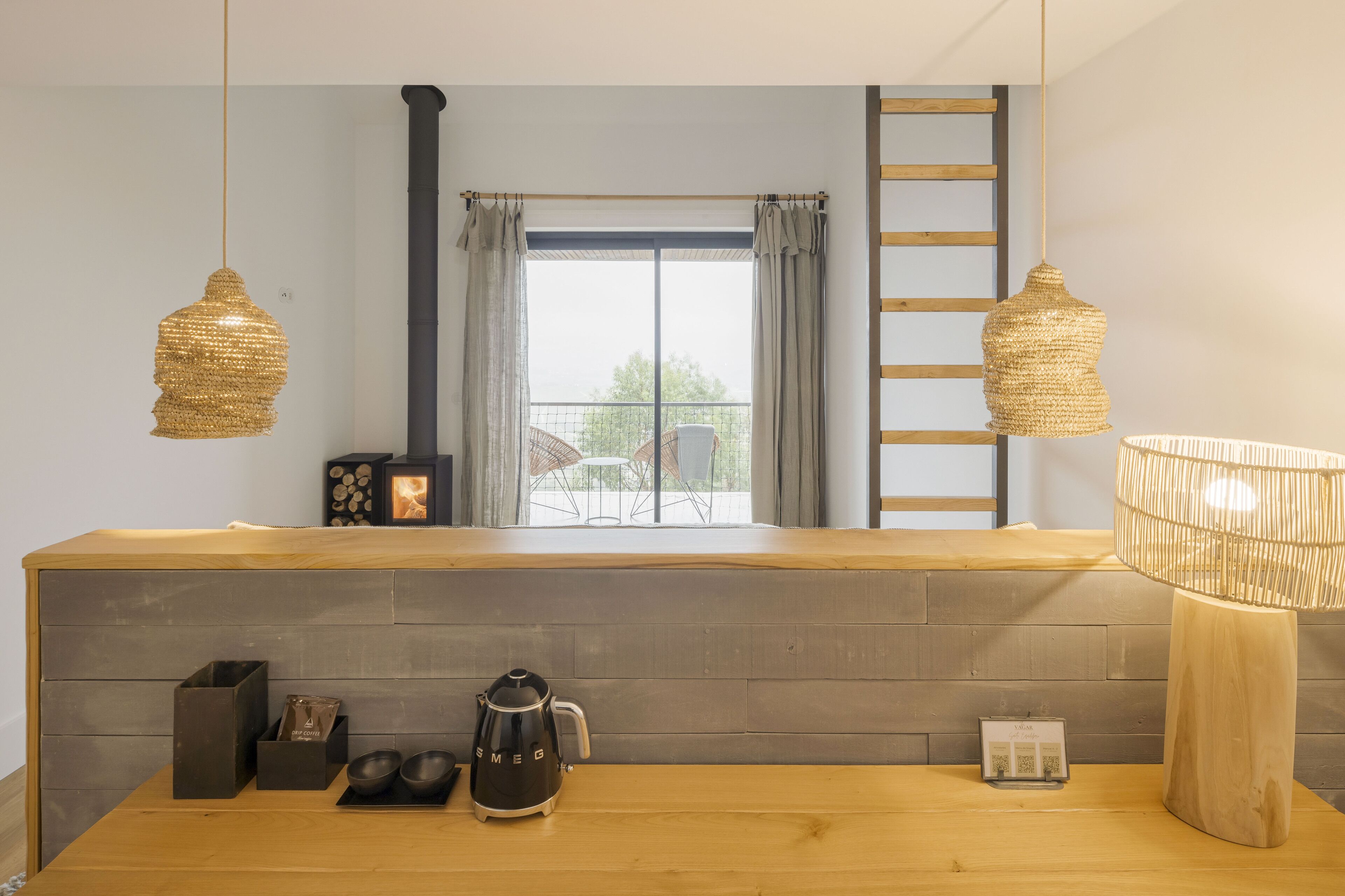 Una cocina moderna con encimera de madera, luminarias tejidas colgantes, una cafetera y vistas al exterior a través de un amplio ventanal.