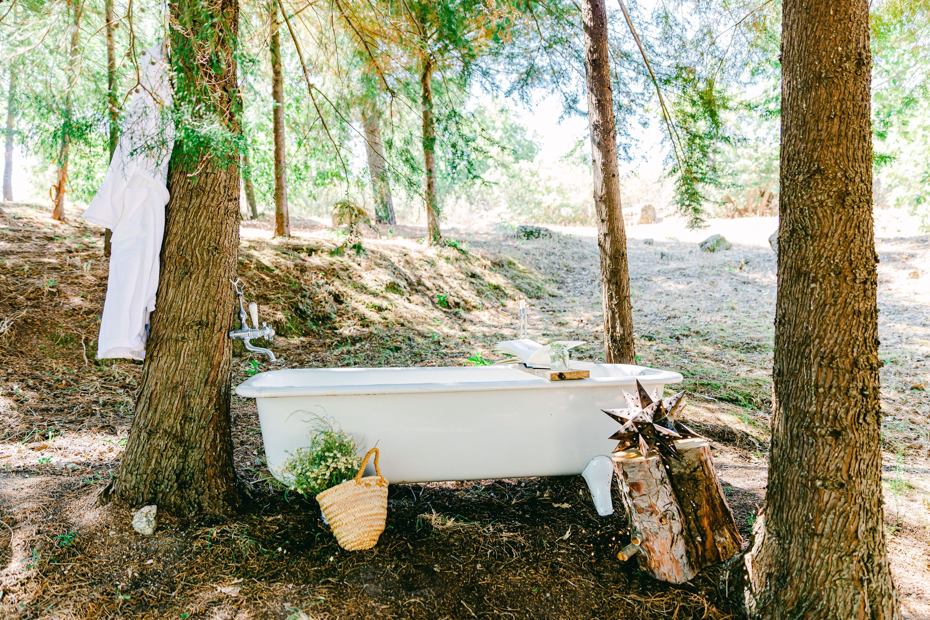 A white bathtub placed outdoors among trees, with a white cloth hanging on one tree and a woven basket on the ground.