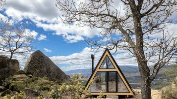 A triangular glass structure with a metal frame, surrounded by rocks and sparse vegetation, overlooking a mountainous landscape.