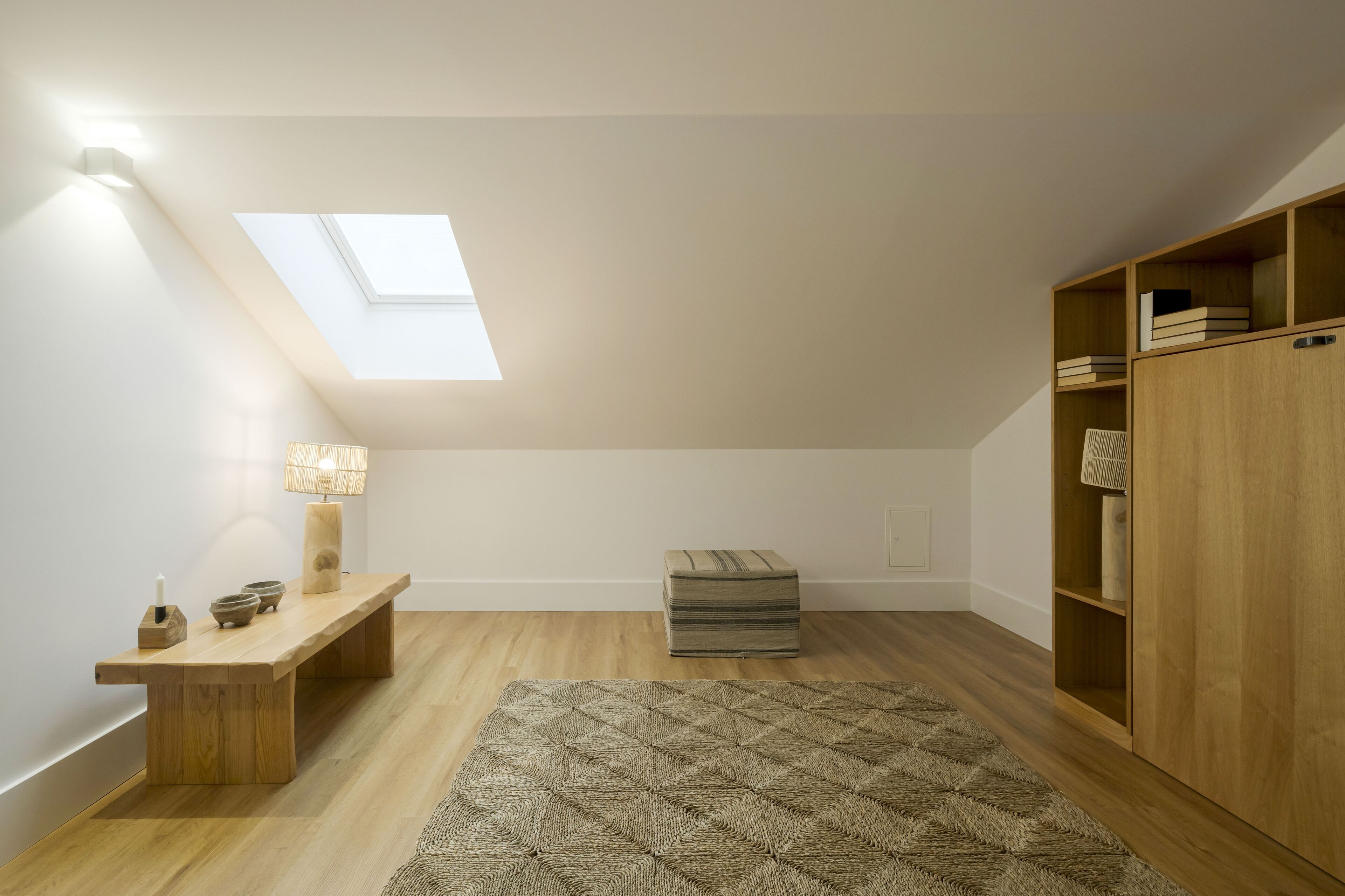 Una habitación con tragaluz, banca de madera, mesa con velas y un tapete estampado.