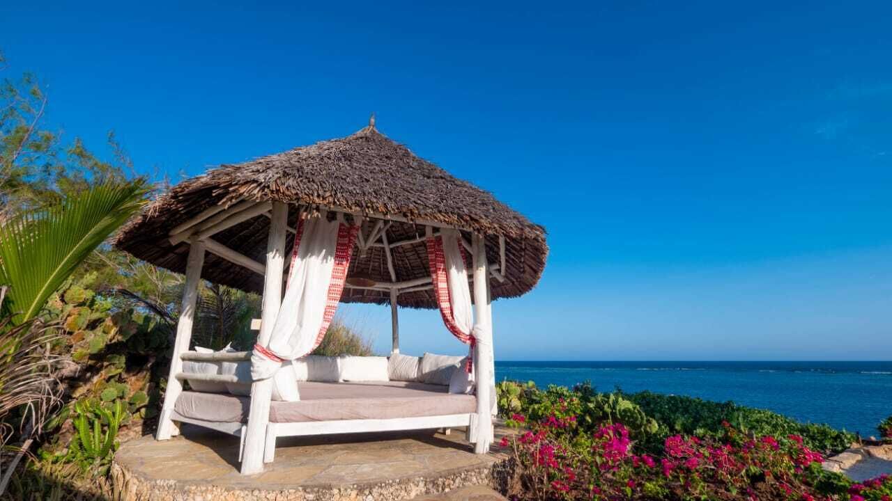 A gazebo with a thatched roof and white pillars, surrounded by lush greenery and a view of the ocean.