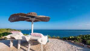 Un espacio de descanso frente a la playa con una sombrilla tejida y sillas blancas acolchadas.