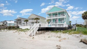 Una propiedad frente a la playa con una casa de dos pisos, un balcón, una terraza de madera y una escalera que baja a la playa.